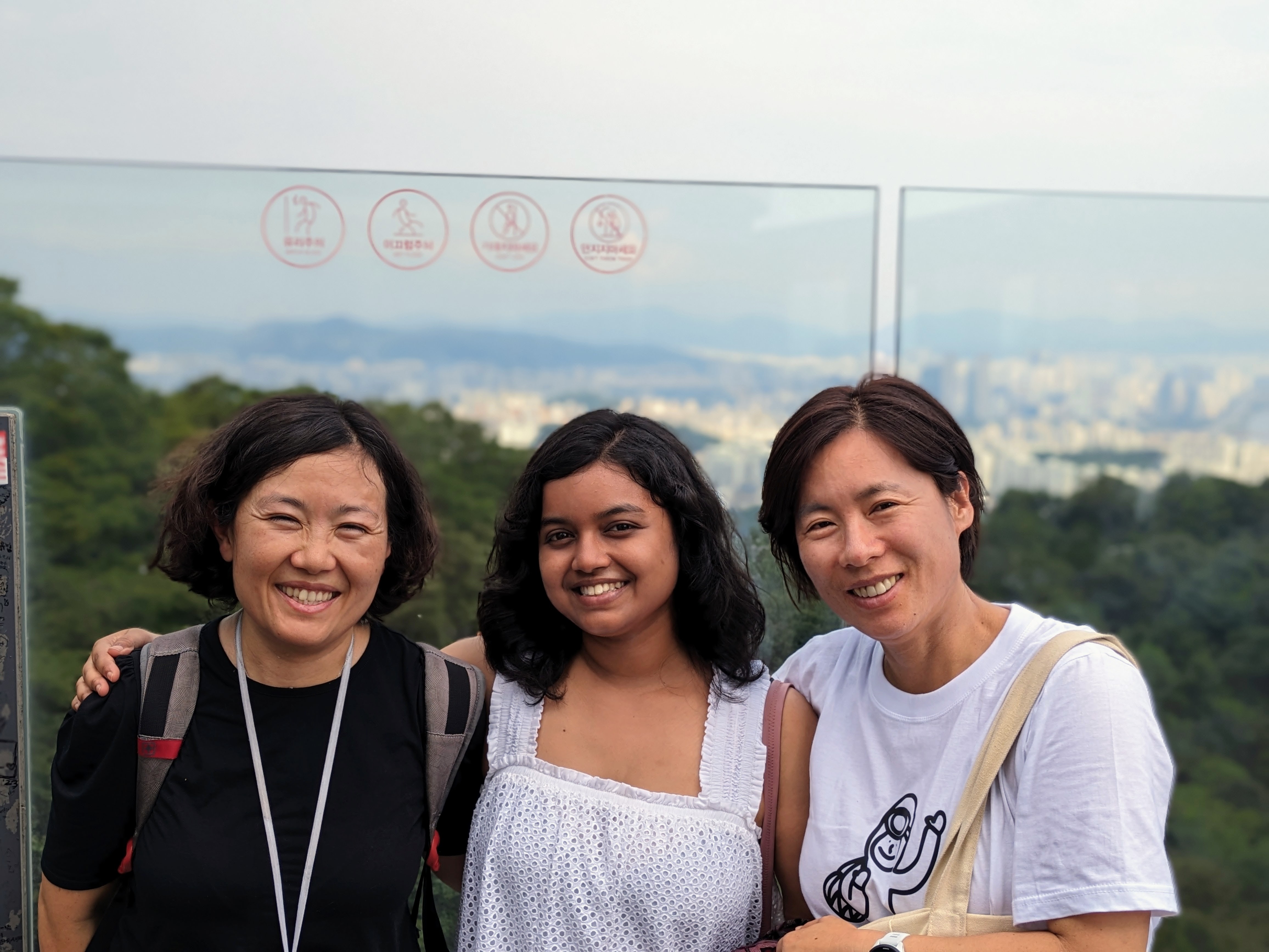 Three smiling women stand close together outdoors at an elevated viewpoint. They are positioned in front of a clear glass safety barrier that overlooks a distant city skyline nestled among rolling green hills under an overcast sky.