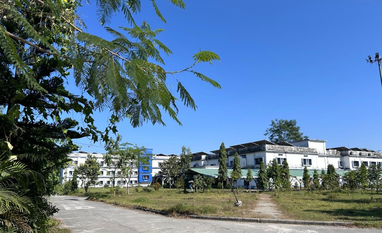 An outdoor shot of a white multi-story building complex with solar panels on the roof under a clear blue sky. A paved path curves in the foreground, bordered by green lawns, small trees, and lush overhanging foliage.