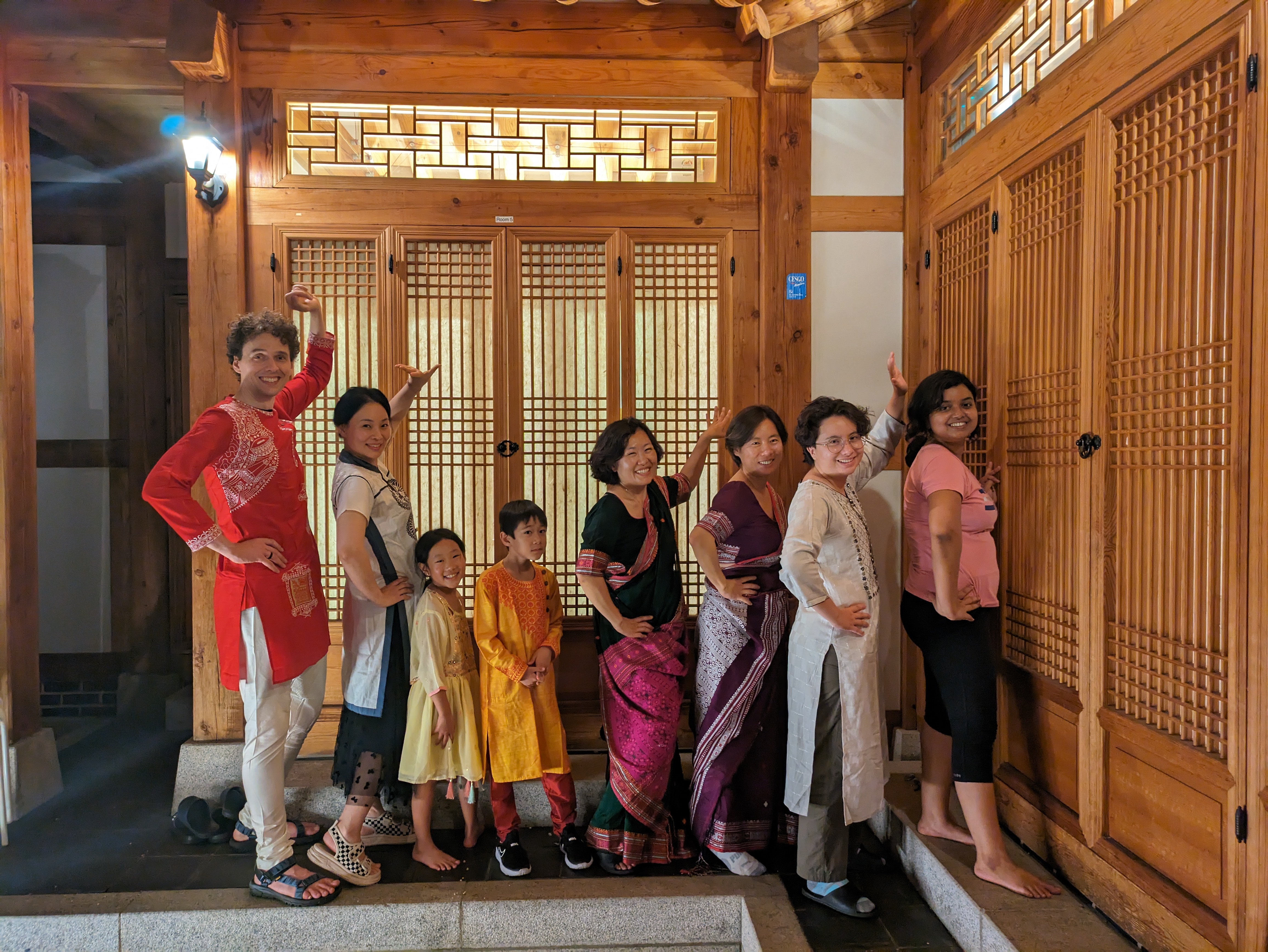 A group of eight people, including two young children, pose playfully in a row outside a traditional building with intricate wooden grid doors and warm lighting. Several individuals are wearing traditional Indian attire, including kurtas and sarees, and many have one hand on their hip or raised in the air.