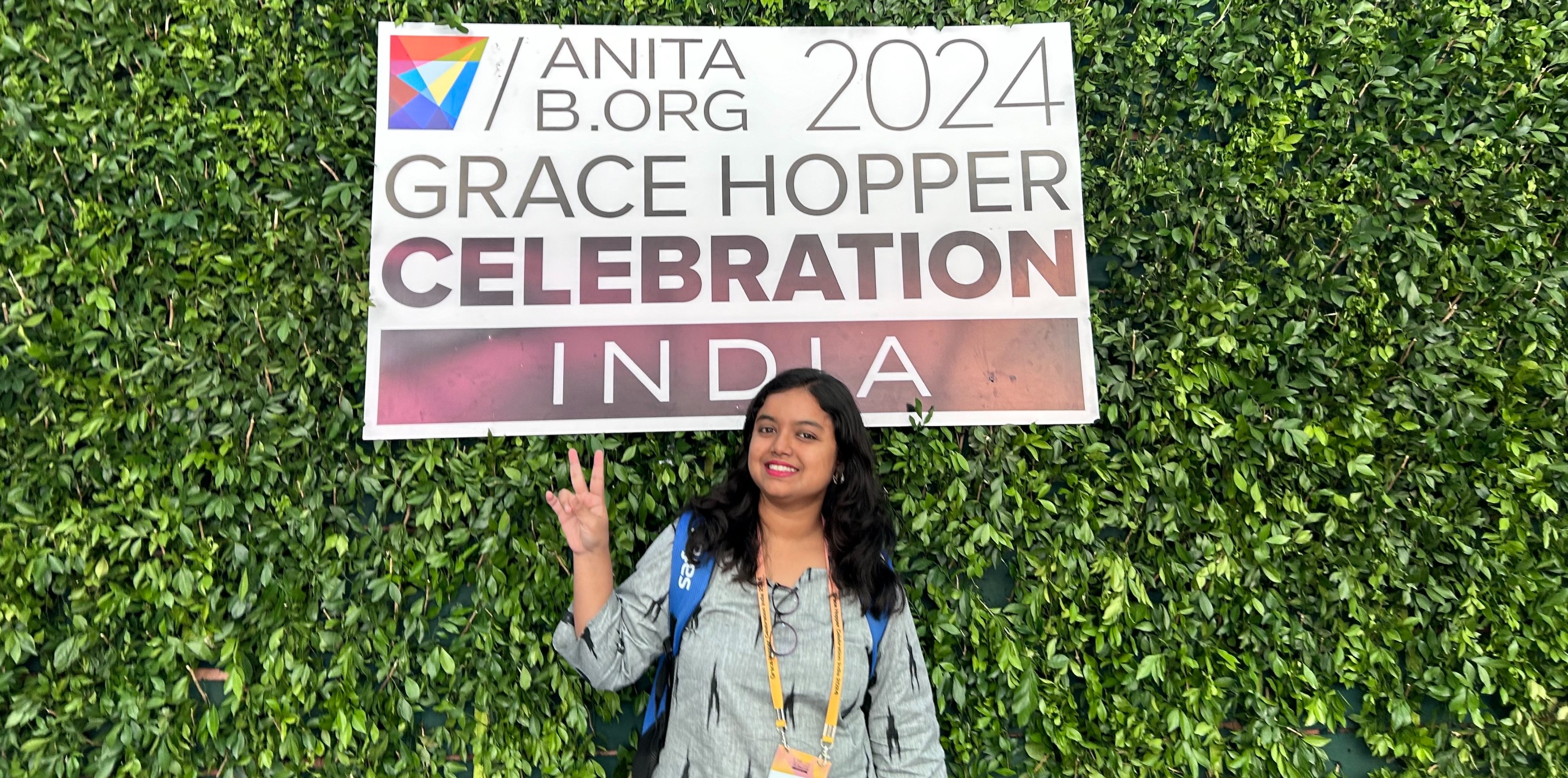 A smiling woman poses and gives a peace sign in front of a green hedge wall. A large sign mounted on the wall reads, 